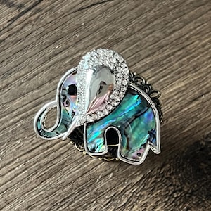 Può includere: Spilla a forma di elefante con dettagli in madreperla iridescente e una cornice in metallo argentato. La testa dell'elefante è adornata con strass trasparenti. La spilla è posta su uno sfondo di legno.