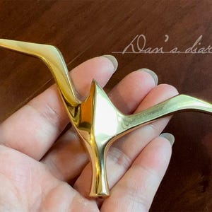 Puede incluir: Un objeto decorativo dorado con forma de pájaro con las alas extendidas. El objeto se sostiene en una mano sobre un fondo marrón. El texto "Dani's diary" está escrito en cursiva en la esquina superior derecha.