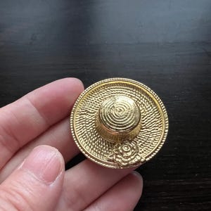 Könnte beinhalten: Eine goldfarbene Metallnadel in Form eines Strohhutes mit einem Blumendetail.