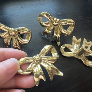 May include: Four gold-colored bow-shaped cabinet pulls. One pull is being held by a hand. The pulls have a shiny finish and are designed to look like tied bows. They are likely used for drawers or cabinets.