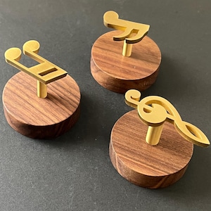 Può includere: Tre pomelli decorativi per mobili con disegni di note musicali. Ogni pomello ha un simbolo musicale color oro montato su una base rotonda in legno. Le basi sono di colore marrone caldo, su sfondo scuro.
