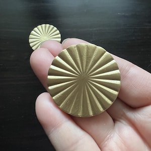 Peut inclure: Deux boutons de meuble circulaires dorés avec un motif de soleil. Les boutons sont en métal et ont une surface texturée. L'un des boutons est tenu dans une main, l'autre est en arrière-plan.