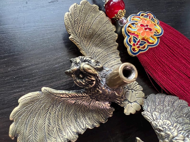 K&ouml;nnte beinhalten: Dekoratives Objekt in Form eines Vogels aus Messing mit ausgebreiteten Fl&uuml;geln. Es hat ein detailliertes Design und eine zylindrische &Ouml;ffnung. Dazu geh&ouml;rt eine rote Quaste mit einem gestickten Blumenemblem. Der Stil ist vintage und dekorativ.