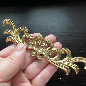 May include: Gold-toned metal decorative trim with a scroll design. The trim has a delicate, intricate pattern with a floral motif.