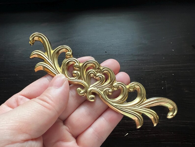 May include: Gold-toned metal decorative trim with a scroll design. The trim has a delicate, intricate pattern with a floral motif.