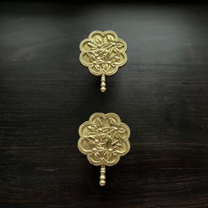 May include: Two gold-toned metal wall hooks with a floral design. Each hook has a round base with a floral pattern and a small, round knob at the bottom.