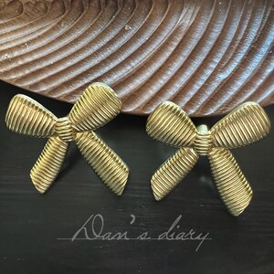 Pode incluir: Um par de brincos dourados em forma de laço. Os brincos têm uma textura canelada e estão sobre um fundo escuro com um detalhe em madeira. O texto "Dan's diary" está escrito à mão.