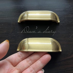 Könnte beinhalten: Zwei goldfarbene Schranktürgriffe mit abgerundeter, leicht geschwungener Form. Die Griffe sind aus Metall und haben eine gebürstete Oberfläche.