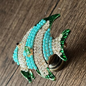 以下が含まれることがあります： ターコイズ、白、緑のアクセントが施された、魚の形をした装飾的なブローチ。 ターコイズビーズとクリアクリスタル、ひれと縁にグリーンのエナメルディテールが特徴です。 魚はゴールドトーンの金属ベースにセットされています。