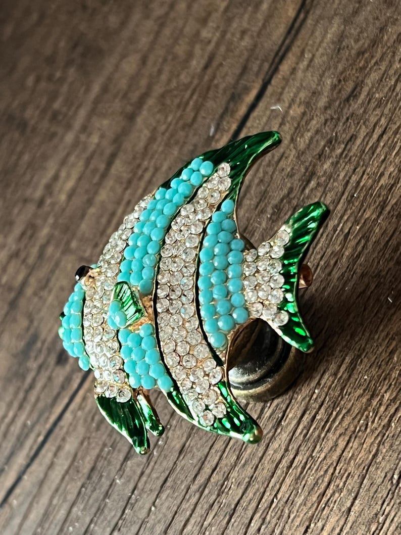 以下が含まれることがあります： ターコイズ、白、緑のアクセントが施された、魚の形をした装飾的なブローチ。 ターコイズビーズとクリアクリスタル、ひれと縁にグリーンのエナメルディテールが特徴です。 魚はゴールドトーンの金属ベースにセットされています。