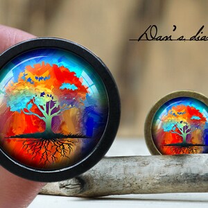May include: Two round, decorative cabochons with a colorful tree design. The larger cabochon has a black bezel, while the smaller one has a bronze bezel. The tree features a green trunk and branches with a vibrant, multi-colored canopy.