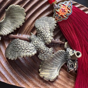 K&ouml;nnte beinhalten: Auf einer geschnitzten Holzoberfl&auml;che liegen verzierte Messingv&ouml;gel mit ausgebreiteten Fl&uuml;geln, m&ouml;glicherweise Flaschenverschl&uuml;sse. Ein roter Quastenanh&auml;nger mit dekorativen Elementen setzt Farbakzente. Die V&ouml;gel weisen detaillierte Federmuster auf.