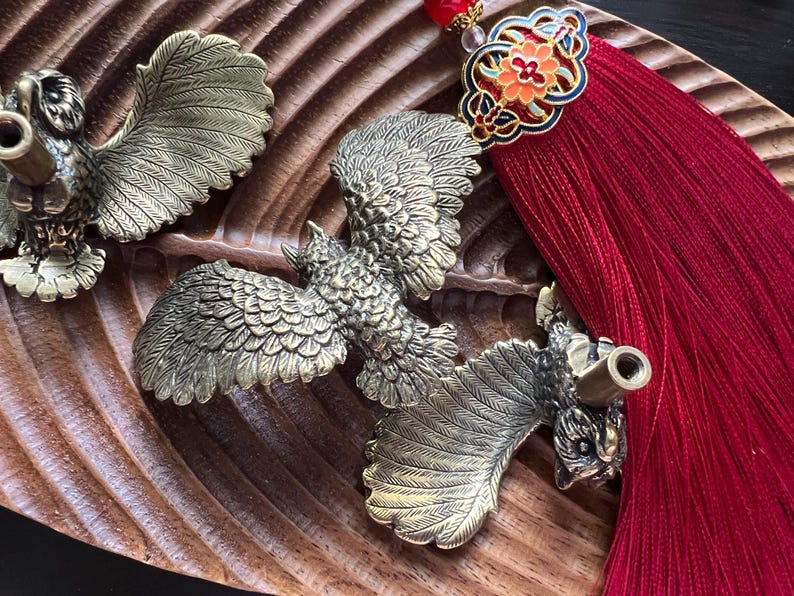 K&ouml;nnte beinhalten: Auf einer geschnitzten Holzoberfl&auml;che liegen verzierte Messingv&ouml;gel mit ausgebreiteten Fl&uuml;geln, m&ouml;glicherweise Flaschenverschl&uuml;sse. Ein roter Quastenanh&auml;nger mit dekorativen Elementen setzt Farbakzente. Die V&ouml;gel weisen detaillierte Federmuster auf.