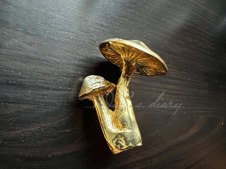 May include: A gold-colored metal sculpture of two mushrooms. The larger mushroom has a wide, flat cap and a slender stem. The smaller mushroom is nestled under the larger one.