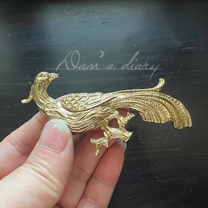 Puede incluir: Un broche dorado con forma de pájaro con detalles intrincados. El broche presenta un diseño de pájaro de cola larga con un cuerpo texturizado y una cola detallada. El texto "Dan's diary" está escrito en cursiva en el fondo.