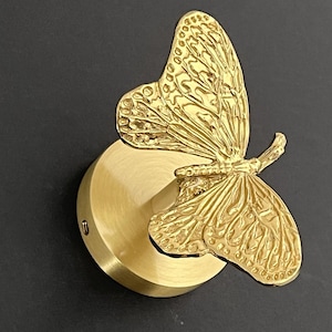 Könnte beinhalten: Goldfarbene Wandleuchte in Schmetterlingsform. Der Schmetterling hat detaillierte Flügelmuster und ist auf einer runden, messingfarbenen Basis montiert. Die Leuchte ist für die Wandmontage konzipiert und wirkt dekorativ.