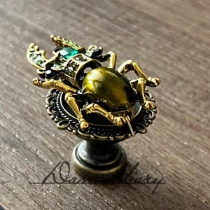 May include: A decorative cabinet knob featuring a detailed beetle design. The beetle has a gold-colored body and legs, with green and black accents. The knob is mounted on a decorative base.
