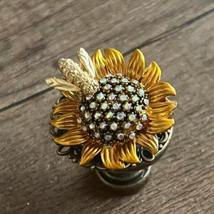 Könnte beinhalten: Ein dekorativer knauf in Sonnenblumenform mit einem Bienenakzent. Die Sonnenblume hat gelbe Blütenblätter und eine Mitte, die mit schillernden Kristallen verziert ist. Die Biene ist goldfarben und der Knauf hat eine verzierte, bronzefarbene Basis.