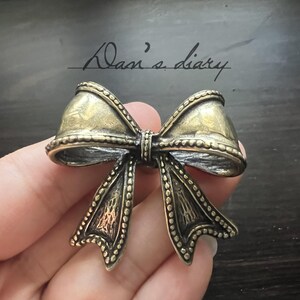 Pode incluir: Broche em forma de laço de estilo vintage em tons de ouro e prata antigos. O broche apresenta detalhes intrincados, incluindo uma borda com contas e superfícies texturizadas. O texto manuscrito "Dan's diary" é visível no fundo.