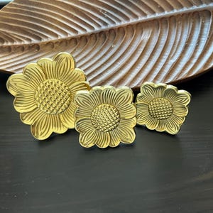 Peut inclure: Trois tirettes de tiroir en métal de couleur dorée en forme de fleur. La plus grande tirette est au centre et les deux plus petites sont de chaque côté.