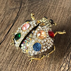 May include: A gold-coloured ladybird brooch adorned with clear rhinestones and colourful gemstones. The brooch features green, pink, orange, red, and blue gemstone accents. The ladybird's body is covered in small, sparkling rhinestones.