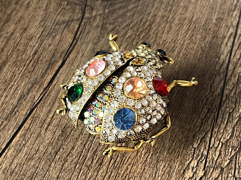 May include: A gold-coloured ladybird brooch adorned with clear rhinestones and colourful gemstones. The brooch features green, pink, orange, red, and blue gemstone accents. The ladybird's body is covered in small, sparkling rhinestones.