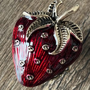 Op de afbeelding: Een decoratieve aardbeivormige sieradendoos. De doos is helderrood met goudkleurige accenten op de bladeren en zaden. De doos met scharnieren is gemaakt van metaal en heeft een gedetailleerd ontwerp.