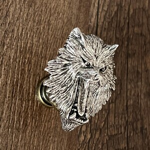 K&ouml;nnte beinhalten: Ein silberfarbener Schrankknauf in Form eines Wolfskopfes mit grimmigem Ausdruck. Der Knauf hat detailliertes Fell und scharfe Z&auml;hne und ist auf einer messingfarbenen Basis montiert. Der Hintergrund ist eine Holzoberfl&auml;che.