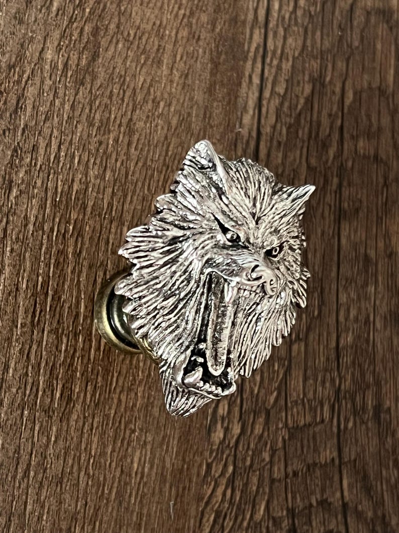 K&ouml;nnte beinhalten: Ein silberfarbener Schrankknauf in Form eines Wolfskopfes mit grimmigem Ausdruck. Der Knauf hat detailliertes Fell und scharfe Z&auml;hne und ist auf einer messingfarbenen Basis montiert. Der Hintergrund ist eine Holzoberfl&auml;che.