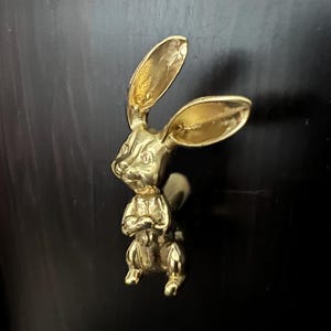 May include: A gold-colored rabbit figurine with long ears. The rabbit is sitting with its arms crossed and has a detailed design.