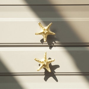 May include: Two gold starfish drawer pulls, mounted on a light grey drawer front.