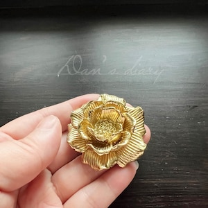 Può includere: Una rosa di metallo dorata con un design dettagliato. La rosa è tenuta in una mano.