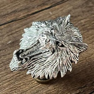 K&ouml;nnte beinhalten: Eine silberfarbene Wolfskopfskulptur mit detailliertem Fell und offenem Maul, das Z&auml;hne zeigt. Die Skulptur ist auf einem kleinen, runden Sockel montiert. Der Hintergrund ist eine Holzoberfl&auml;che.