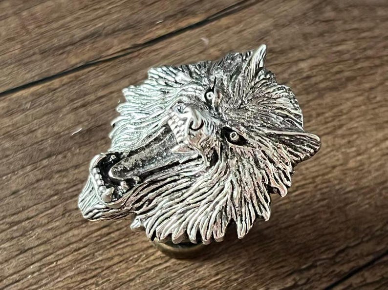 K&ouml;nnte beinhalten: Eine silberfarbene Wolfskopfskulptur mit detailliertem Fell und offenem Maul, das Z&auml;hne zeigt. Die Skulptur ist auf einem kleinen, runden Sockel montiert. Der Hintergrund ist eine Holzoberfl&auml;che.