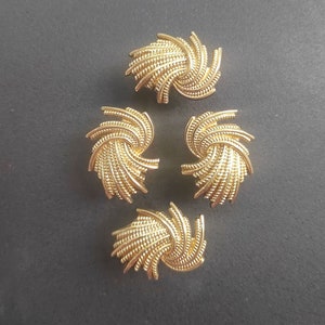 K&ouml;nnte beinhalten: Vier goldfarbene Kn&ouml;pfe mit einem Spiraldesign. Jeder Knopf hat ein strukturiertes, gewebtes Aussehen, mit einem zentralen Wirbel, der sich nach au&szlig;en ausdehnt. Die Kn&ouml;pfe sind auf einer dunkelgrauen Oberfl&auml;che angeordnet, die ihre Details hervorhebt.