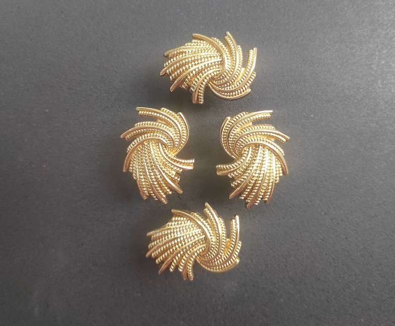 K&ouml;nnte beinhalten: Vier goldfarbene Kn&ouml;pfe mit einem Spiraldesign. Jeder Knopf hat ein strukturiertes, gewebtes Aussehen, mit einem zentralen Wirbel, der sich nach au&szlig;en ausdehnt. Die Kn&ouml;pfe sind auf einer dunkelgrauen Oberfl&auml;che angeordnet, die ihre Details hervorhebt.
