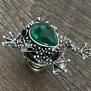 以下が含まれることがあります： 大きなエメラルドグリーンの涙型の石が付いた、銀色のカエルのブローチ。カエルは足が細かく、体がテクスチャ加工されています。上部には黒い石がセットされています。ユニークなアクセサリーです。