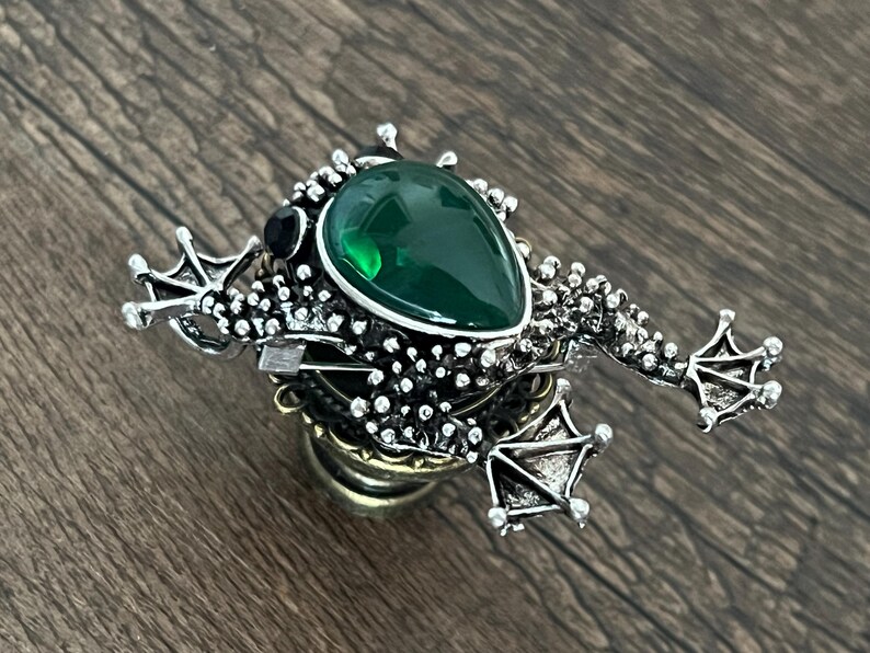 以下が含まれることがあります： 大きなエメラルドグリーンの涙型の石が付いた、銀色のカエルのブローチ。カエルは足が細かく、体がテクスチャ加工されています。上部には黒い石がセットされています。ユニークなアクセサリーです。