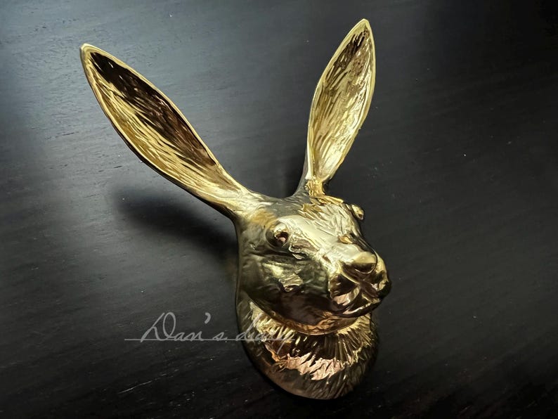 K&ouml;nnte beinhalten: Eine goldfarbene Metallskulptur eines Kaninchenkopfes. Die Skulptur ist detailliert und realistisch, mit strukturiertem Fell und einem lebensechten Ausdruck.