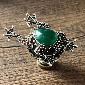 以下が含まれることがあります： 大きな楕円形の緑色の宝石が中央にある、銀色のカエル型のブローチ。ブローチは詳細なテクスチャと小さな足が特徴です。カエルは木製の背景に配置されています。長さ約5cm。