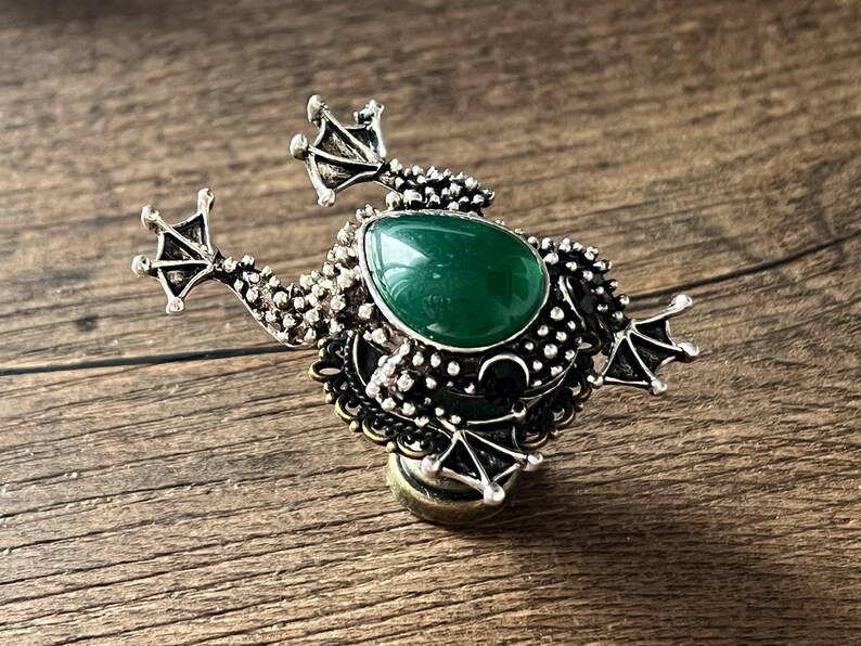 以下が含まれることがあります： 大きな楕円形の緑色の宝石が中央にある、銀色のカエル型のブローチ。ブローチは詳細なテクスチャと小さな足が特徴です。カエルは木製の背景に配置されています。長さ約5cm。