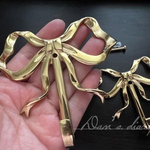 May include: Two gold-coloured metal bow-shaped decorative hooks. The hooks have a detailed ribbon design with a central stem and mounting hole. A screw is visible near the larger hook. The hooks are likely for decorative purposes.