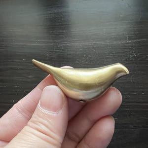 K&ouml;nnte beinhalten: Eine goldfarbene Vogelfigur aus Metall mit einer glatten, abgerundeten Form. Der Vogel sitzt auf einer Hand, mit dem Schnabel nach rechts zeigend.