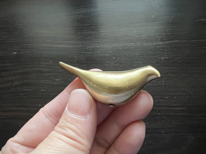 K&ouml;nnte beinhalten: Eine goldfarbene Vogelfigur aus Metall mit einer glatten, abgerundeten Form. Der Vogel sitzt auf einer Hand, mit dem Schnabel nach rechts zeigend.