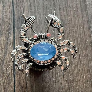 Peut inclure: Broche argentée en forme de crabe, ornée d'une grande pierre ovale bleu clair. Le corps et les pinces du crabe sont ornés de cristaux transparents et roses. La broche est posée sur un fond en bois.