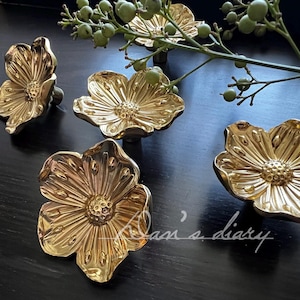 May include: Gold-colored flower-shaped cabinet knobs. The knobs are arranged on a dark surface, with a sprig of greenery in the background. The knobs have a detailed, textured design, resembling a flower with multiple petals.