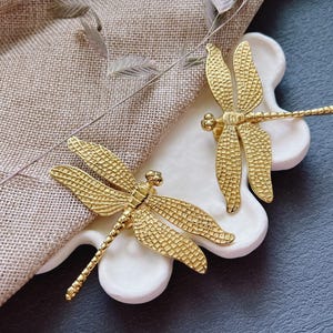 May include: Two gold-toned dragonfly-shaped decorative pieces on a white surface. The dragonflies have detailed wings and bodies.