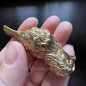 Puede incluir: Una figurita de p&aacute;jaro de metal de color dorado con plumas detalladas. El p&aacute;jaro est&aacute; acostado de lado con la cabeza girada hacia la derecha.