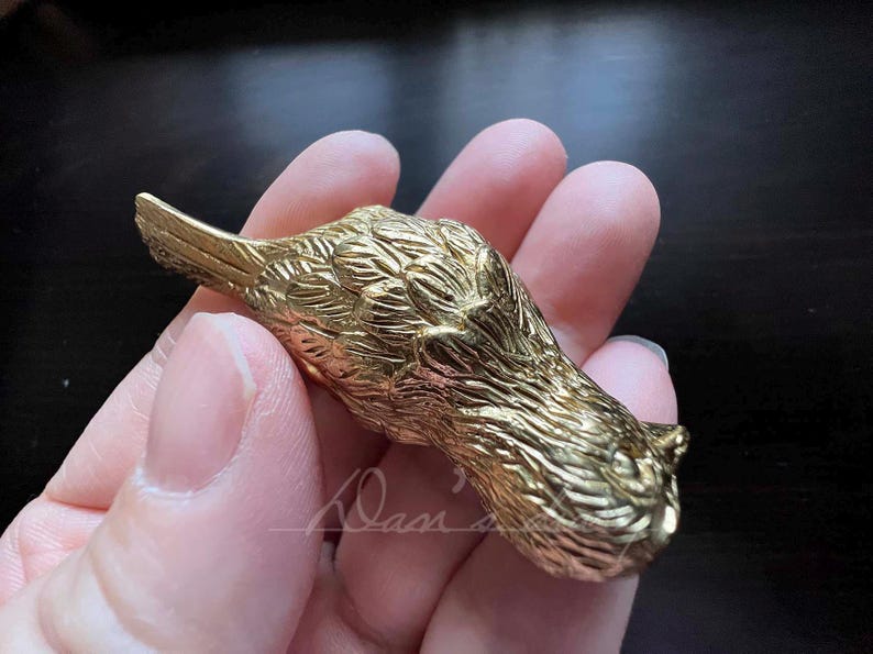 Puede incluir: Una figurita de p&aacute;jaro de metal de color dorado con plumas detalladas. El p&aacute;jaro est&aacute; acostado de lado con la cabeza girada hacia la derecha.