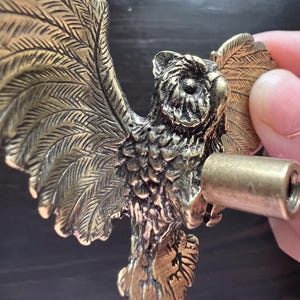 K&ouml;nnte beinhalten: Ein detaillierter, antik-messingfarbener Schrankgriff in Eulenform. Die Eule hat ausgebreitete Fl&uuml;gel mit detaillierten Federn. Ein zylindrischer, messingfarbener Schraubpfosten wird in der N&auml;he des K&ouml;rpers der Eule gehalten, bereit zur Installation. Die Hardware ist f&uuml;r M&ouml;bel konzipiert.